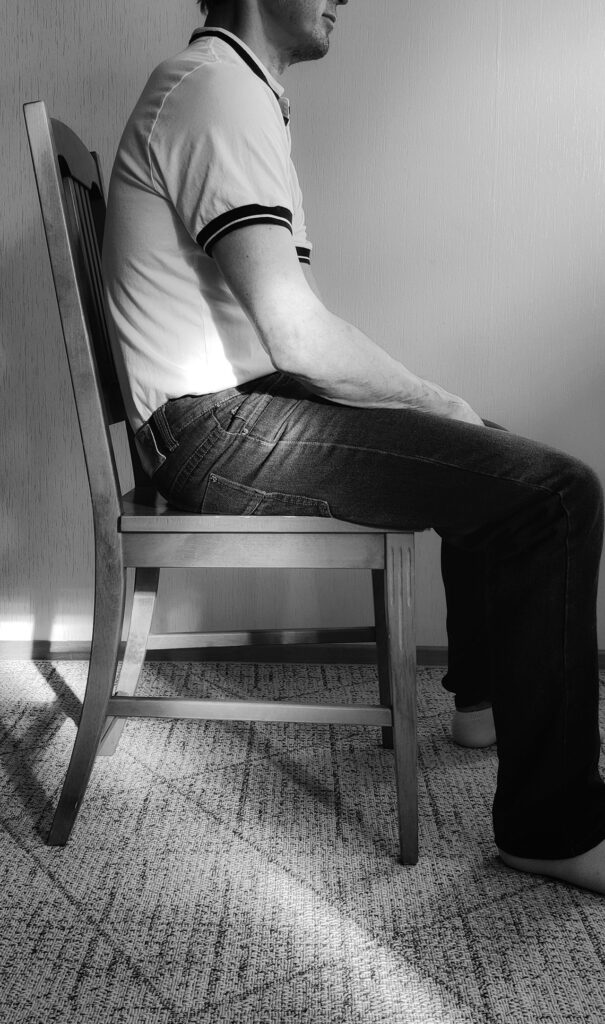 Mies istuu puisella tuolilla hartiat kyyryssä ja selkäranka kaartuu kaarella.